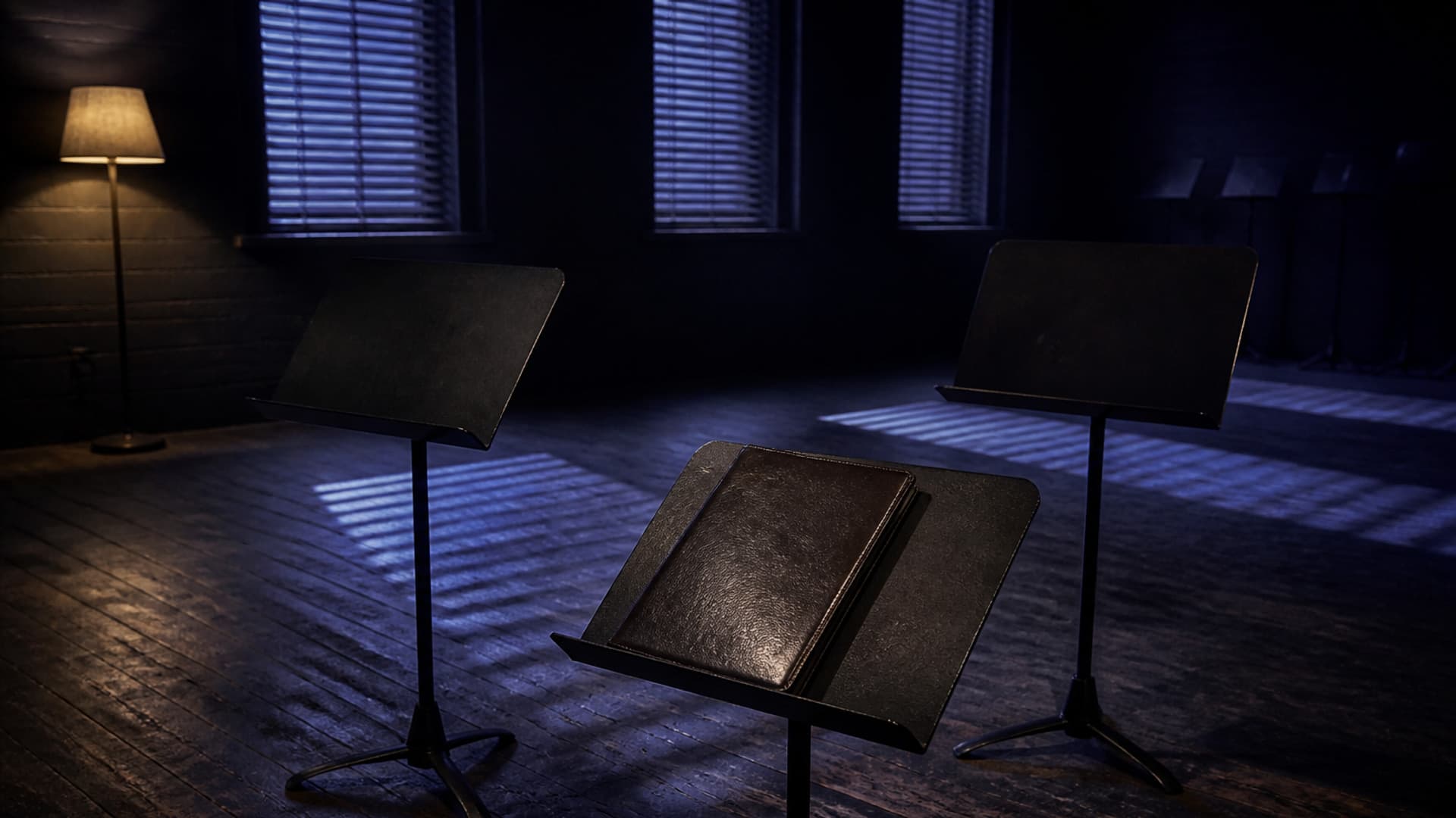 Three empty black music stands on a worn wood floor, a closed unmarked leather folder on the nearest stand, with slatted blue light and no people or readable text