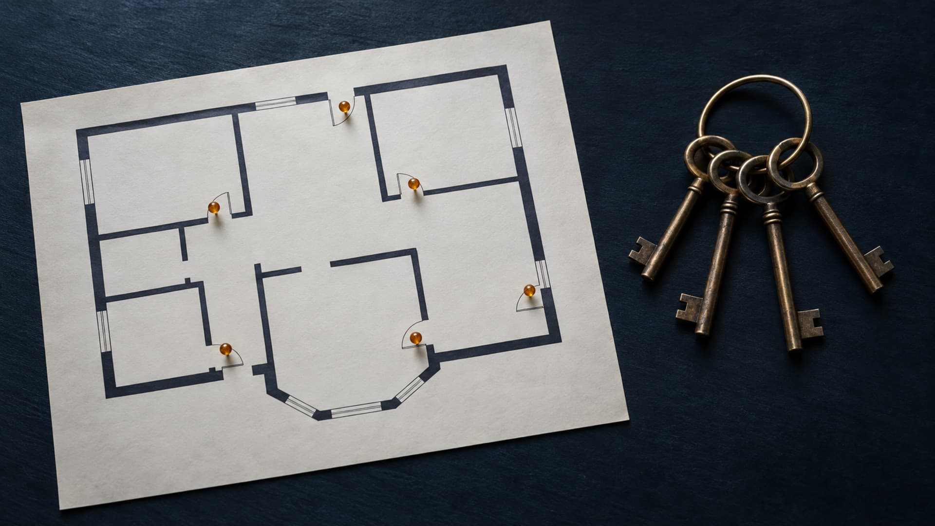 A top-down view of a dark desk with a paper floor plan of a building, several small doorways marked with soft amber pins and one ring of keys resting beside it